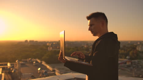Middle-plan:-a-male-freelancer-typing-on-a-laptop-keyboard-at-sunset-overlooking-the-city-while-standing-on-the-roof-of-a-skyscraper.-The-businessman-works-remotely-and-controls-the-work-of-the-company